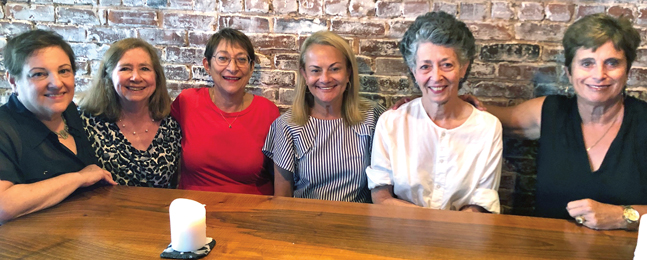 Six smiling women sit behind a long wooden table against a rough brick wall.